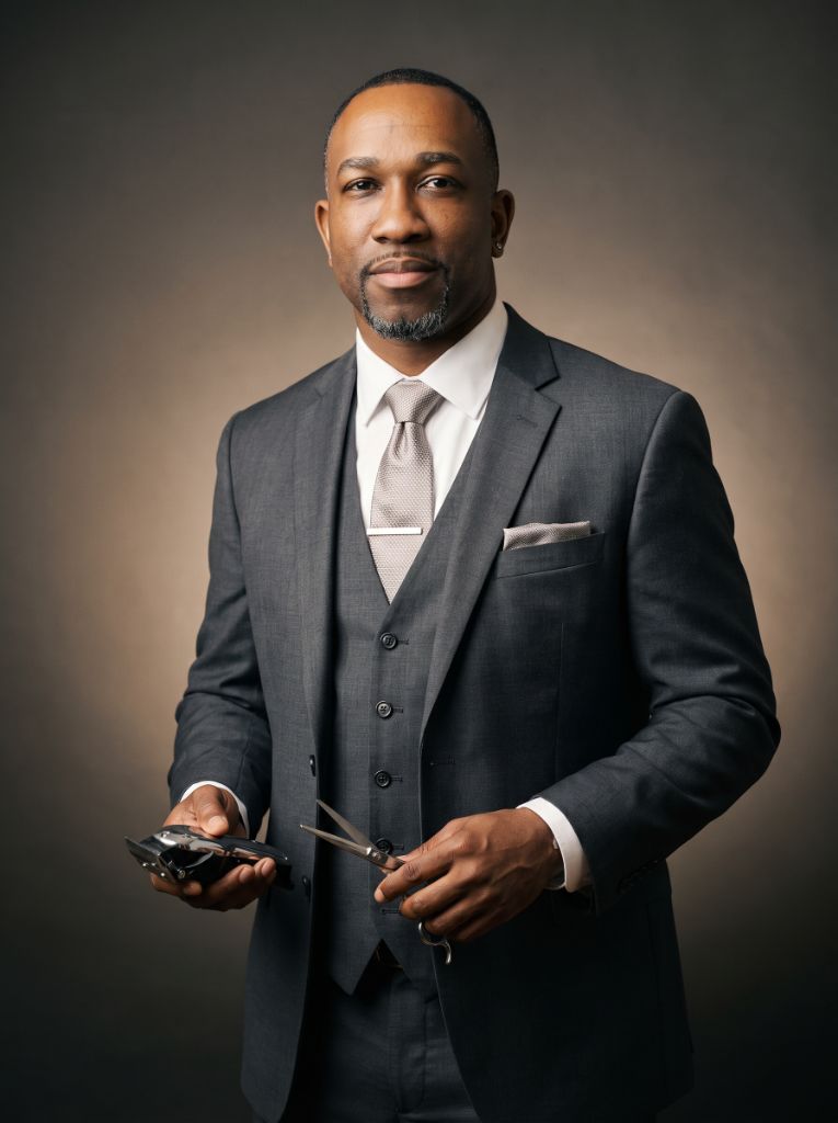 Kelvin Jacob, Founder and Master Barber of The Prestigious Cut, in a charcoal three-piece suit, holding clippers and shears.